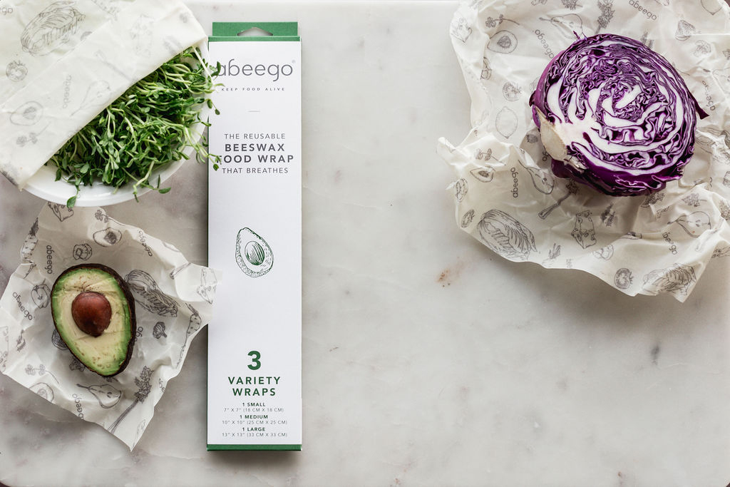 Comptoir de cuisine en marbre blanc avec un bol d'herbes fra&icirc;ches envelopp&eacute; dans Abeego, partiellement d&eacute;couvert ainsi qu'un avocat et un chou violet sur le dessus non emball&eacute; mais couch&eacute; dans un emballage alimentaire en cire Abeego